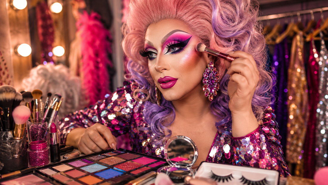 Drag queen aplicando maquillaje con brochas, paleta de sombras, pestañas y accesorios brillantes en una escena colorida de preparación glam.